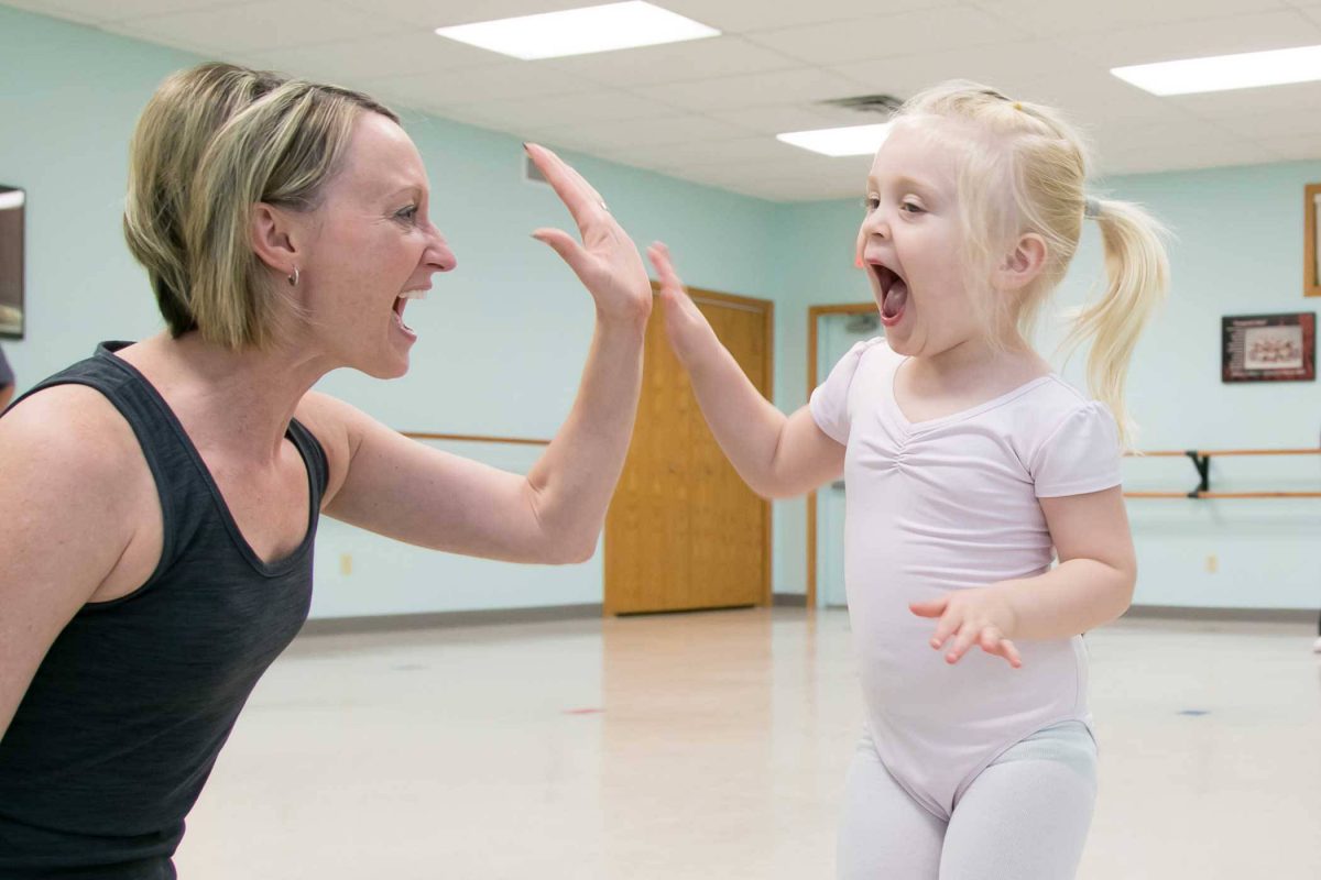 alt="Young, Caucasian blond girl gives dance teacher a high five during toddler dance class"